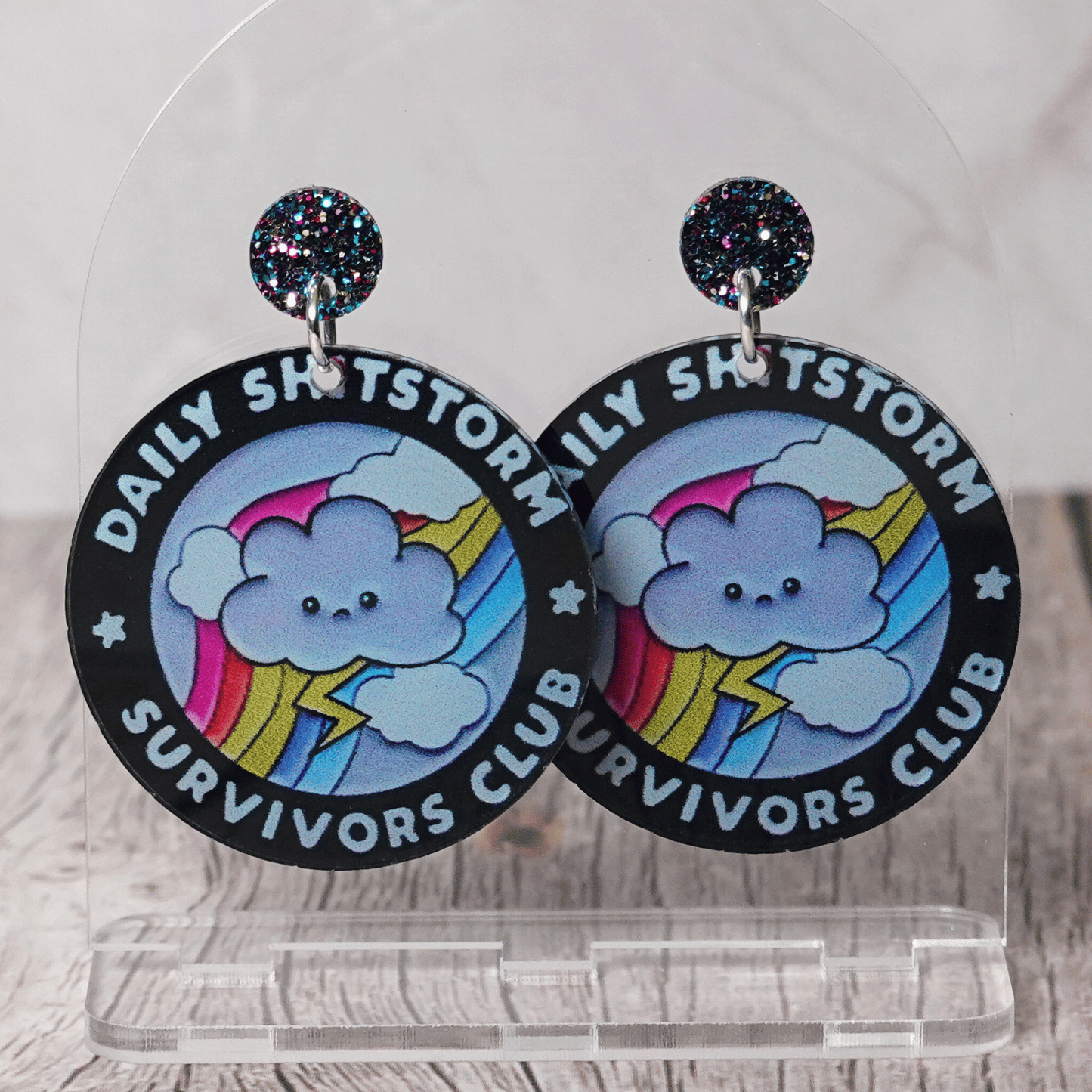 Round novelty dangle earrings reading “Daily Shitstorm Survivors Club” with a cute cloud, rainbow and lightning design