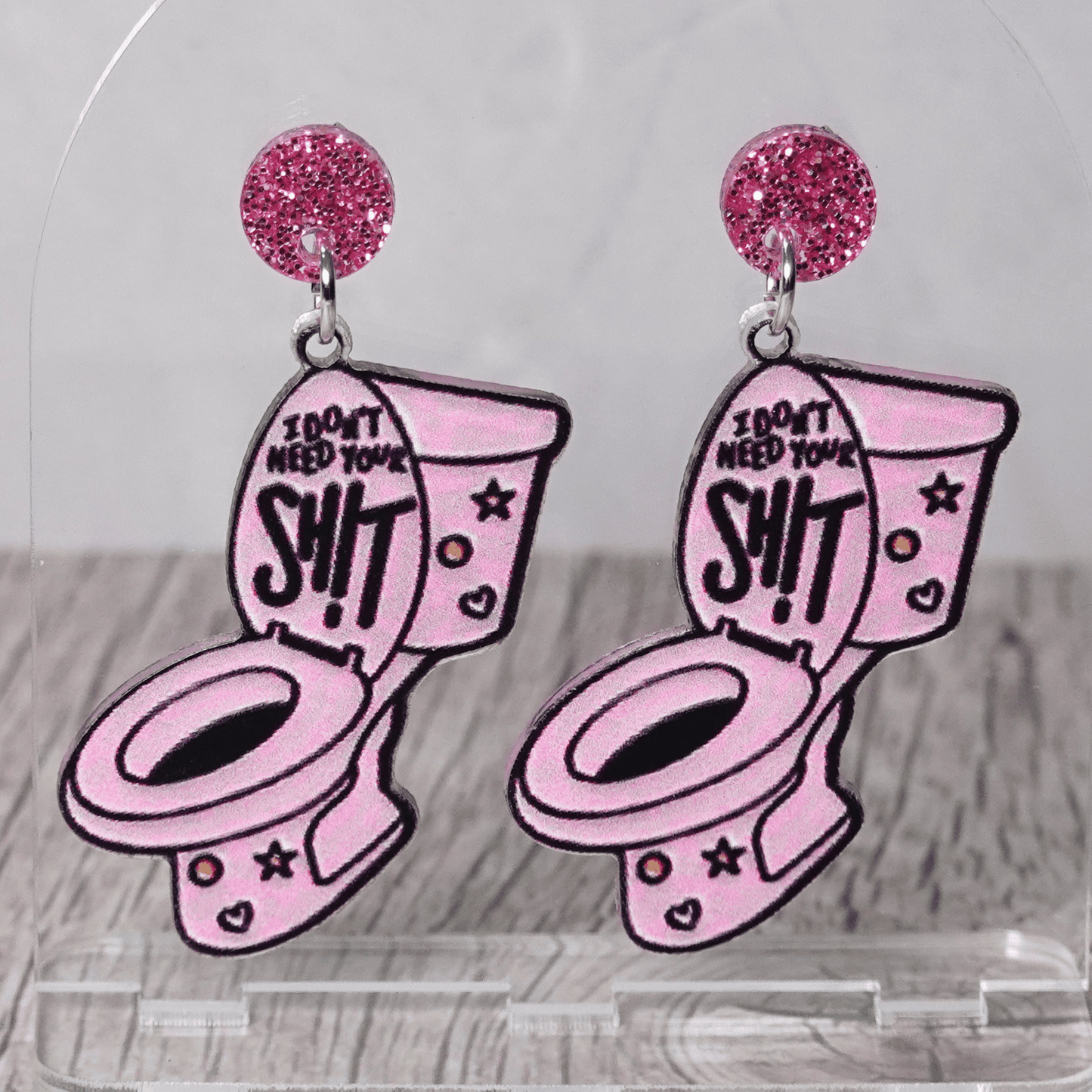 Pink novelty dangle earrings shaped like a toilet with text “I don’t need your sh*t” and glitter stud tops