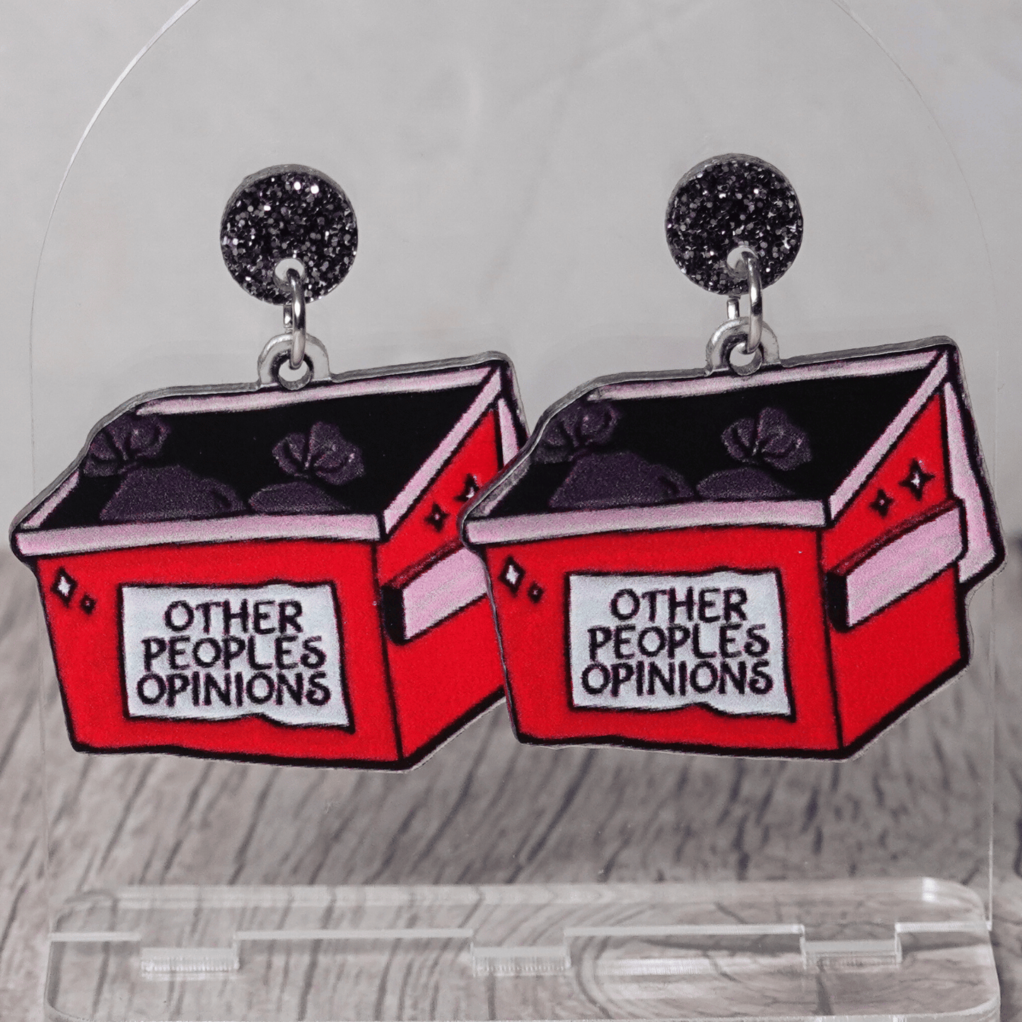 Acrylic dangle earrings shaped like a red dumpster labeled “Other People’s Opinions” with trash bags inside