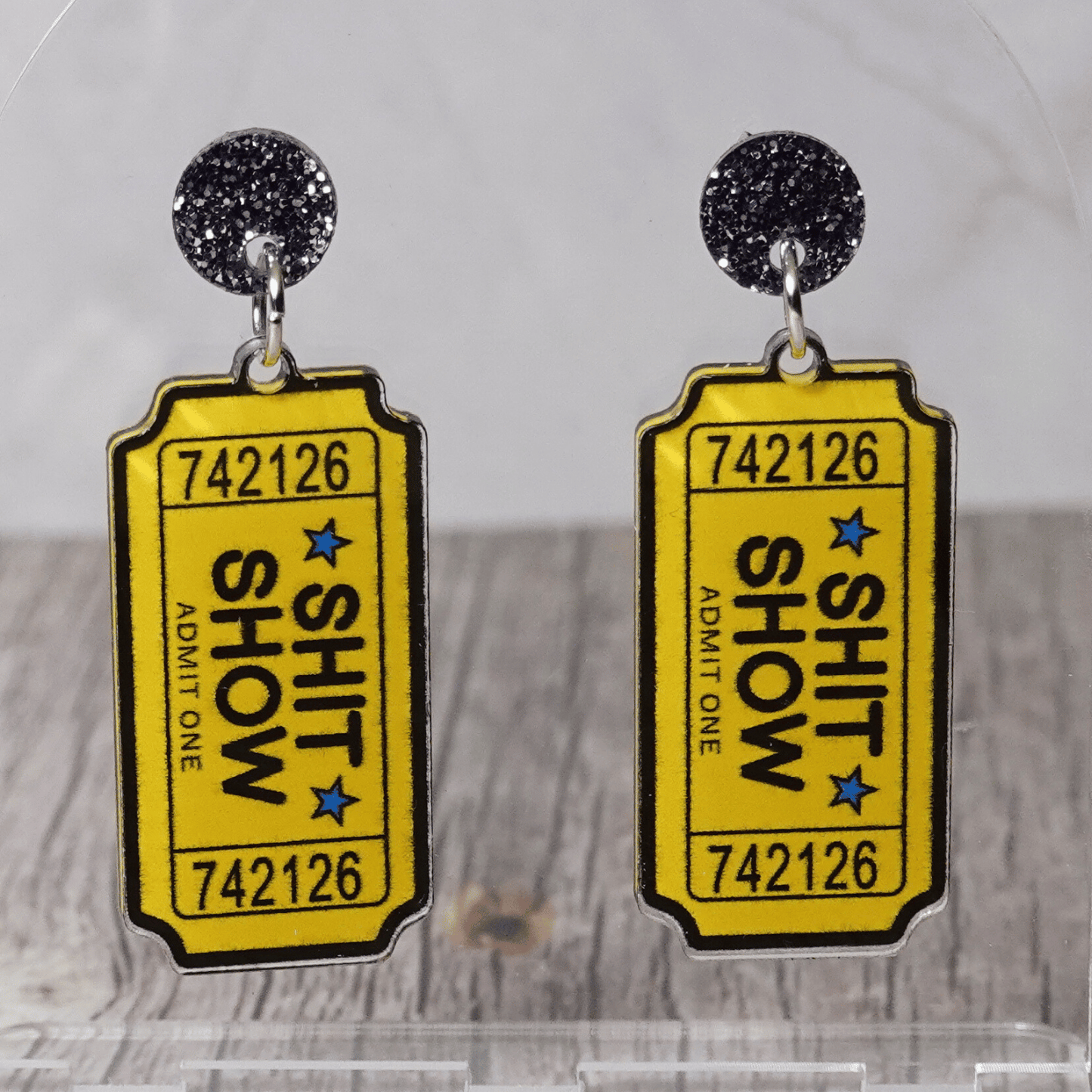 Acrylic dangle earrings designed like yellow admit-one tickets reading “Shit Show”