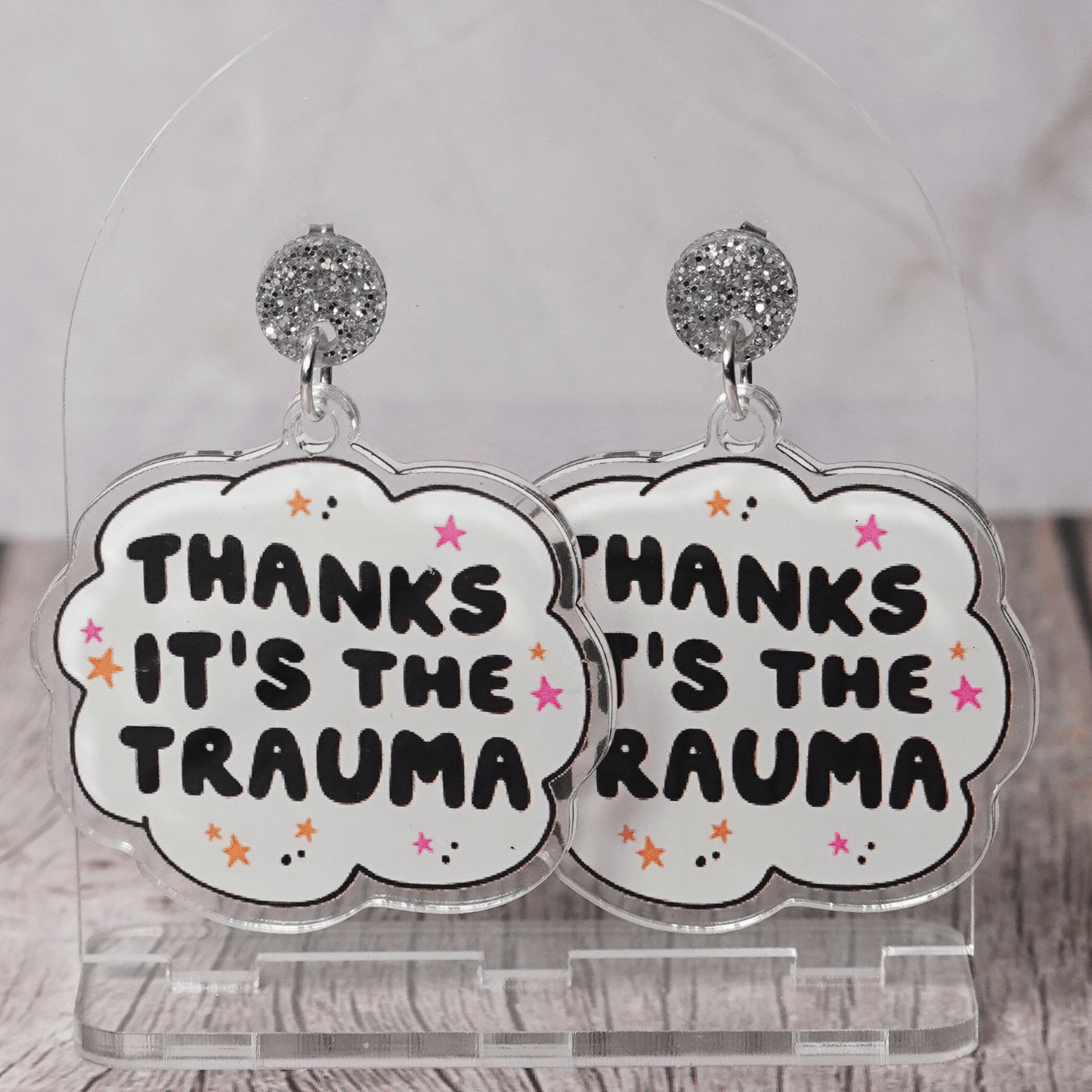 Clear acrylic dangle earrings with a speech bubble shape reading “Thanks it’s the trauma” with small star accents