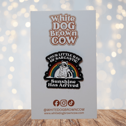 Enamel pin featuring a raccoon in front of a rainbow with the quote “Your little ray of sarcastic sunshine has arrived.”