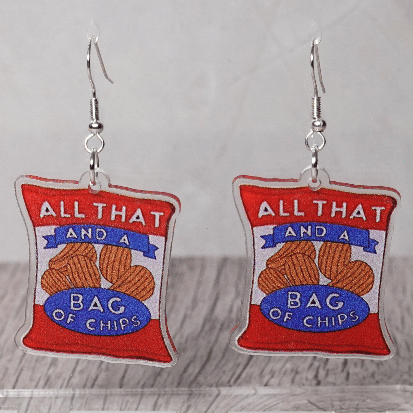 Acrylic dangle earrings shaped like a red potato chip bag that reads “All That and a Bag of Chips,” featuring illustrated chips and bold retro lettering.