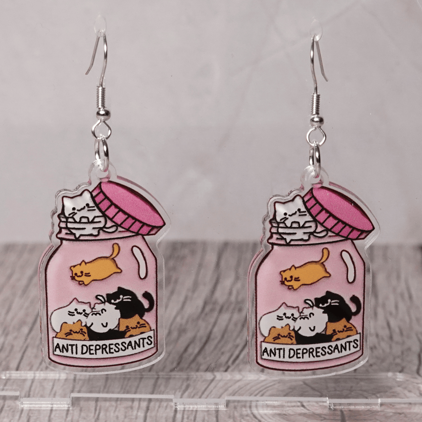 Acrylic dangle earrings shaped like a pink jar labeled “anti-depressants” filled with cartoon cats.