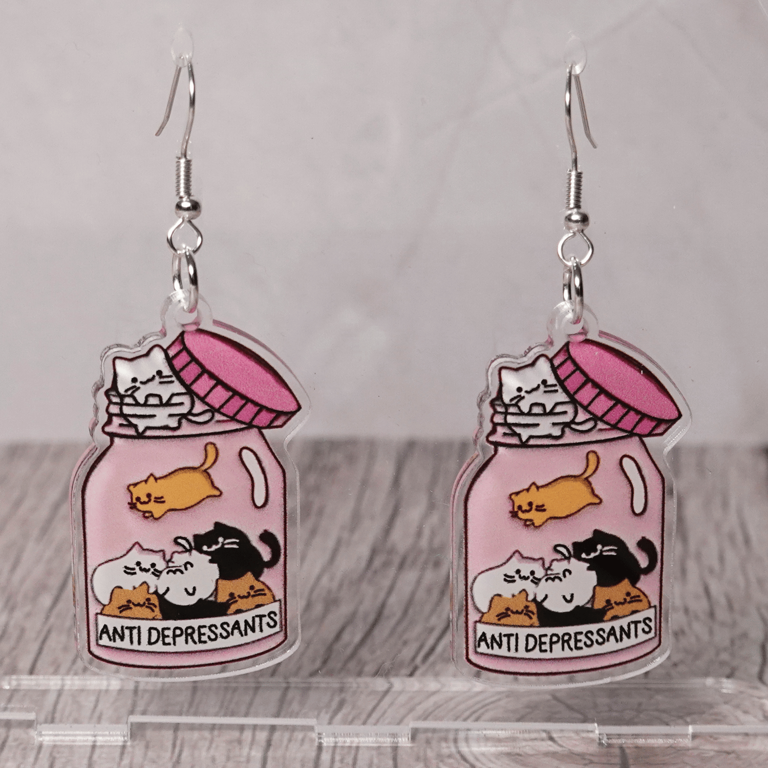Acrylic dangle earrings shaped like a pink jar labeled “anti-depressants” filled with cartoon cats.