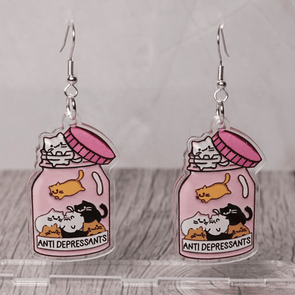 Acrylic dangle earrings shaped like a pink jar labeled “anti-depressants” filled with cartoon cats.