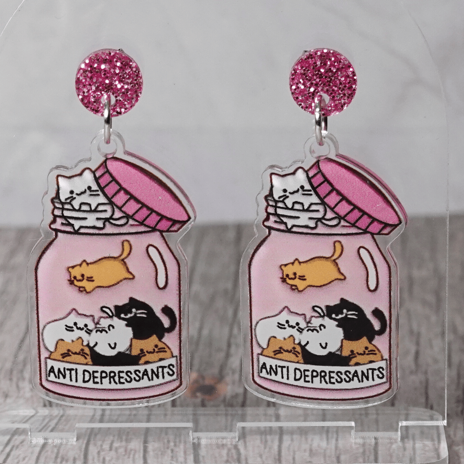 Acrylic dangle earrings shaped like a pink jar labeled “anti-depressants” filled with cartoon cats.