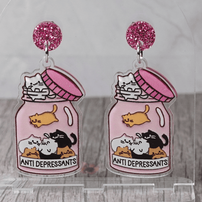Acrylic dangle earrings shaped like a pink jar labeled “anti-depressants” filled with cartoon cats.