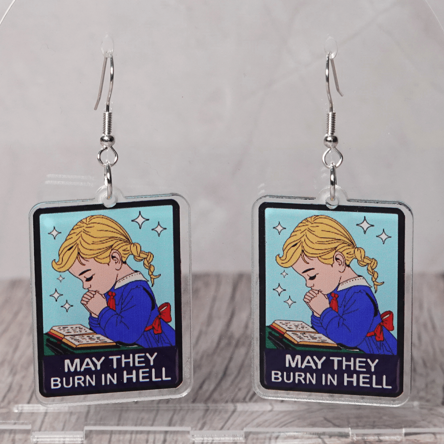 Clear acrylic dangle earrings with a vintage illustration of a praying girl and text reading “May they burn in hell”