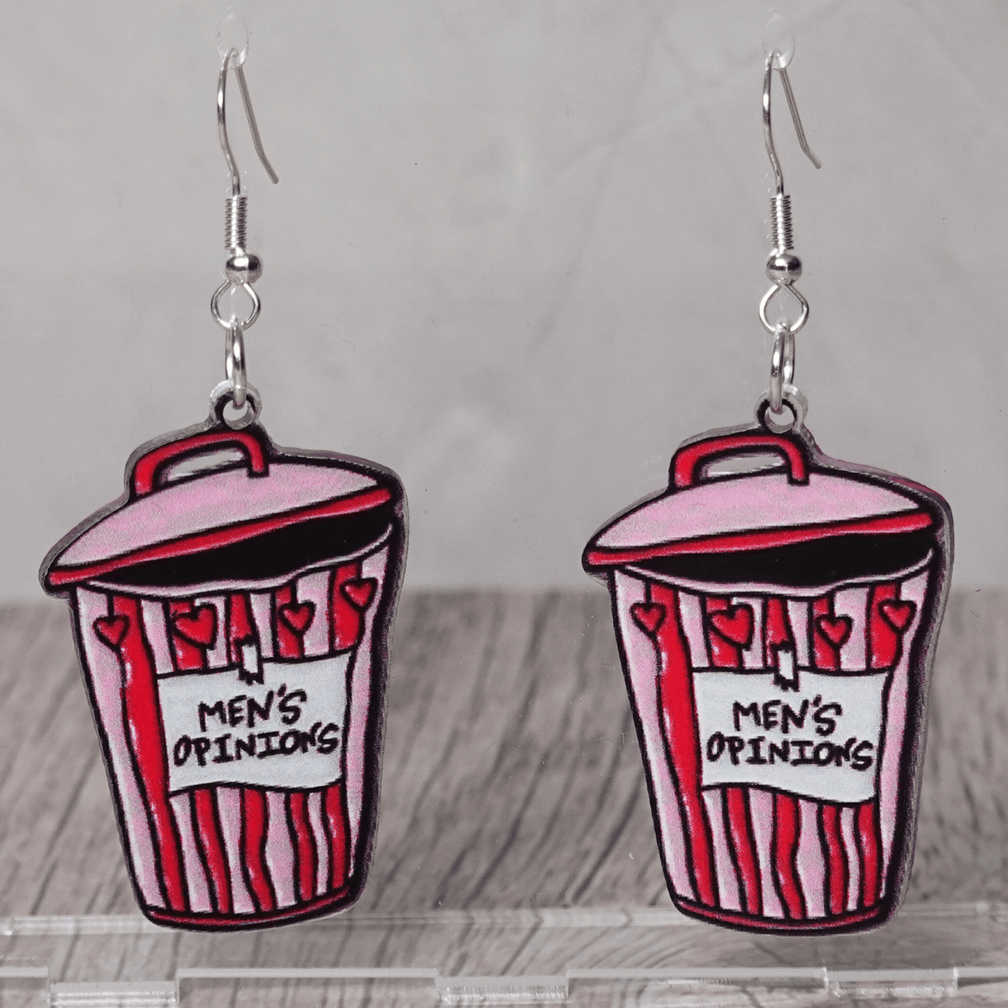 Acrylic dangle earrings shaped like a pink and red trash can labeled “Men’s Opinions”