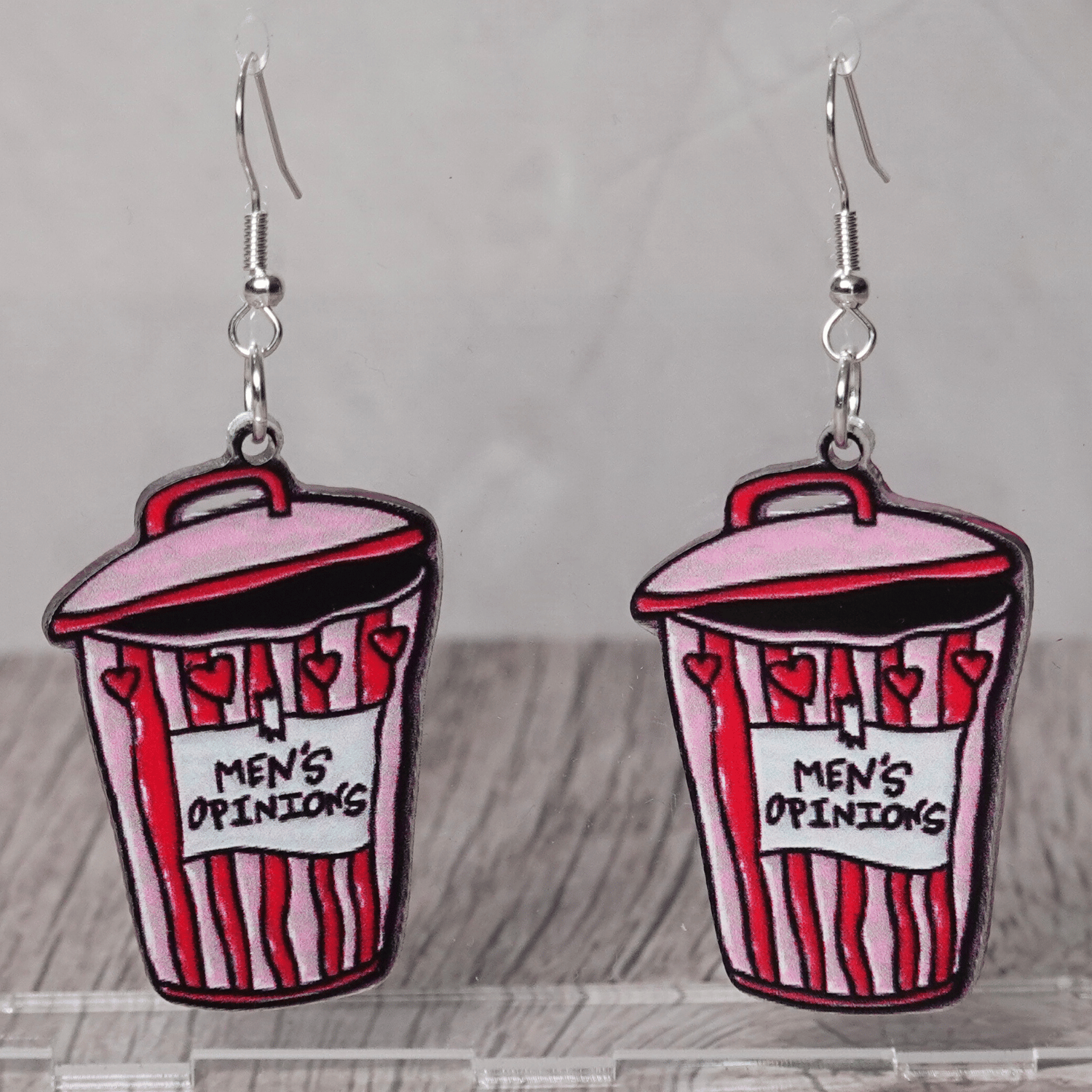 Acrylic dangle earrings shaped like a pink and red trash can labeled “Men’s Opinions”