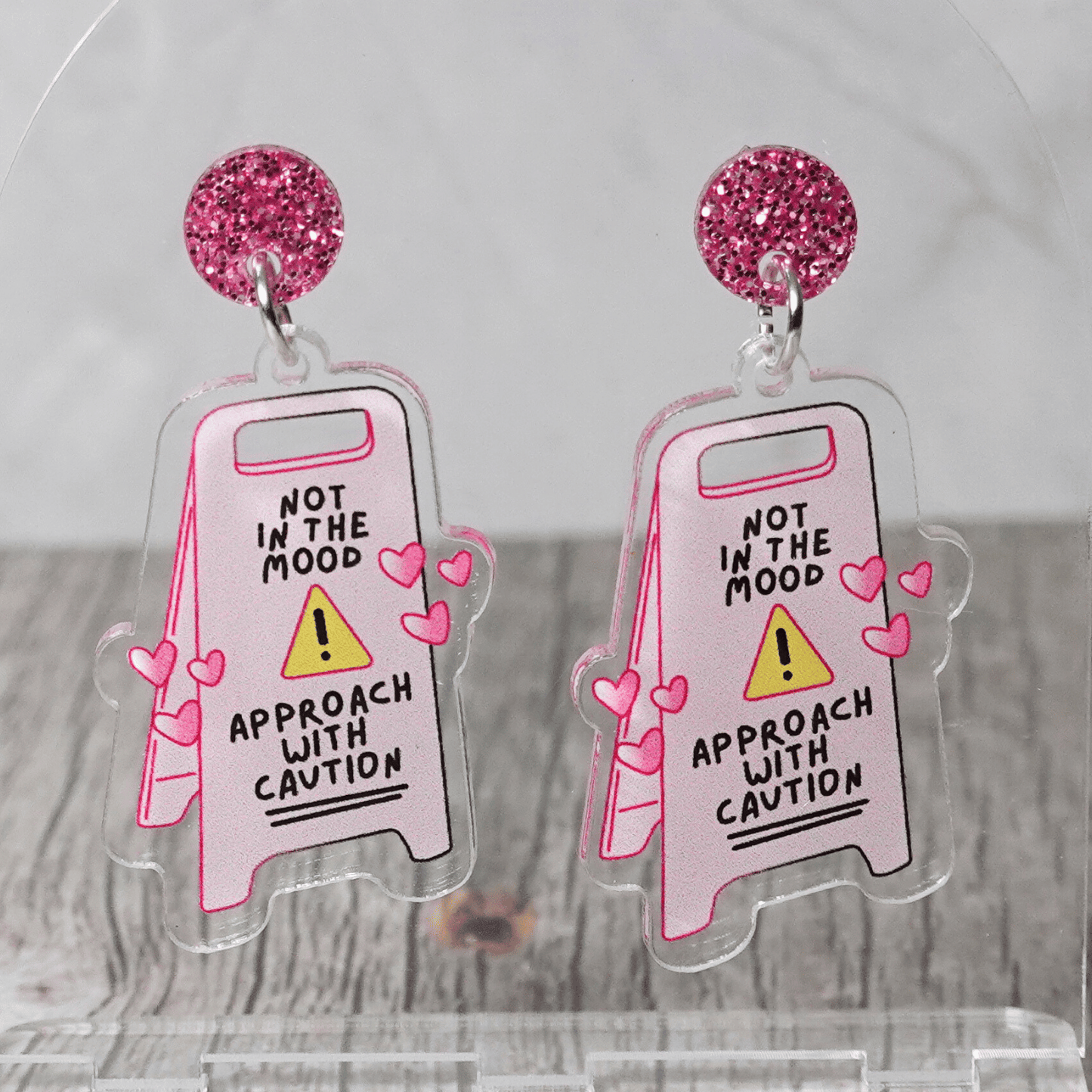 Funny acrylic dangle earrings shaped like a caution sign that reads “Not in the mood – approach with caution,” decorated with small pink hearts.