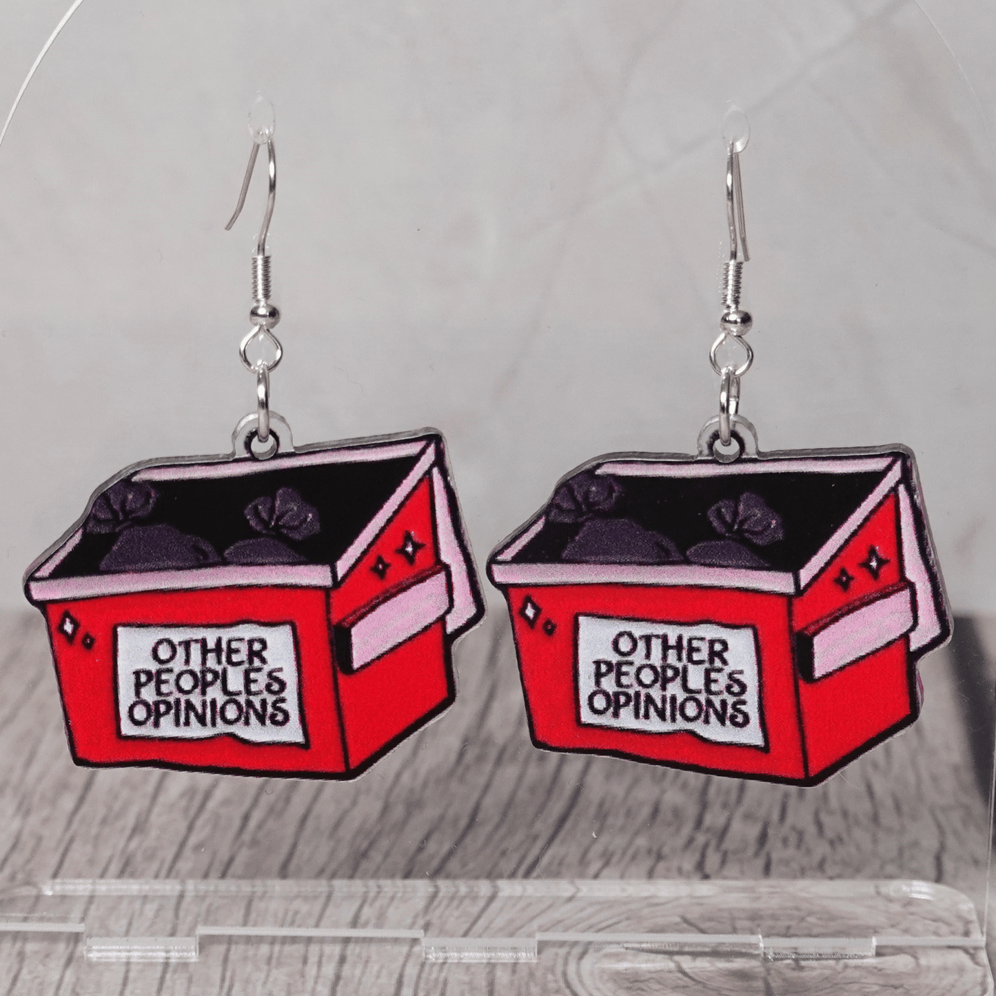 Acrylic dangle earrings shaped like a red dumpster labeled “Other People’s Opinions” with trash bags inside