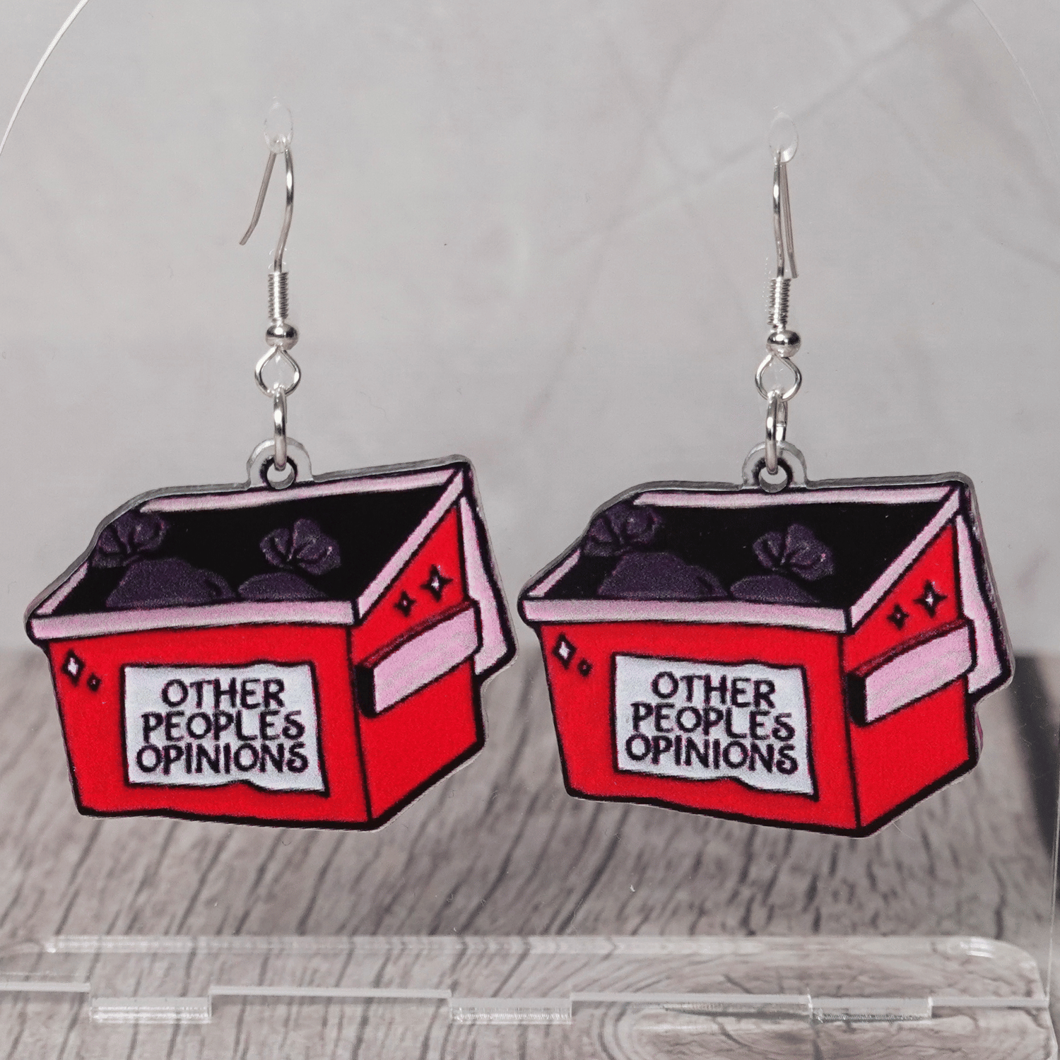 Acrylic dangle earrings shaped like a red dumpster labeled “Other People’s Opinions” with trash bags inside