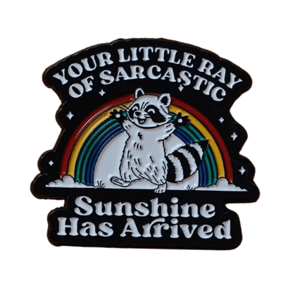 Enamel pin featuring a raccoon in front of a rainbow with the quote “Your little ray of sarcastic sunshine has arrived.”