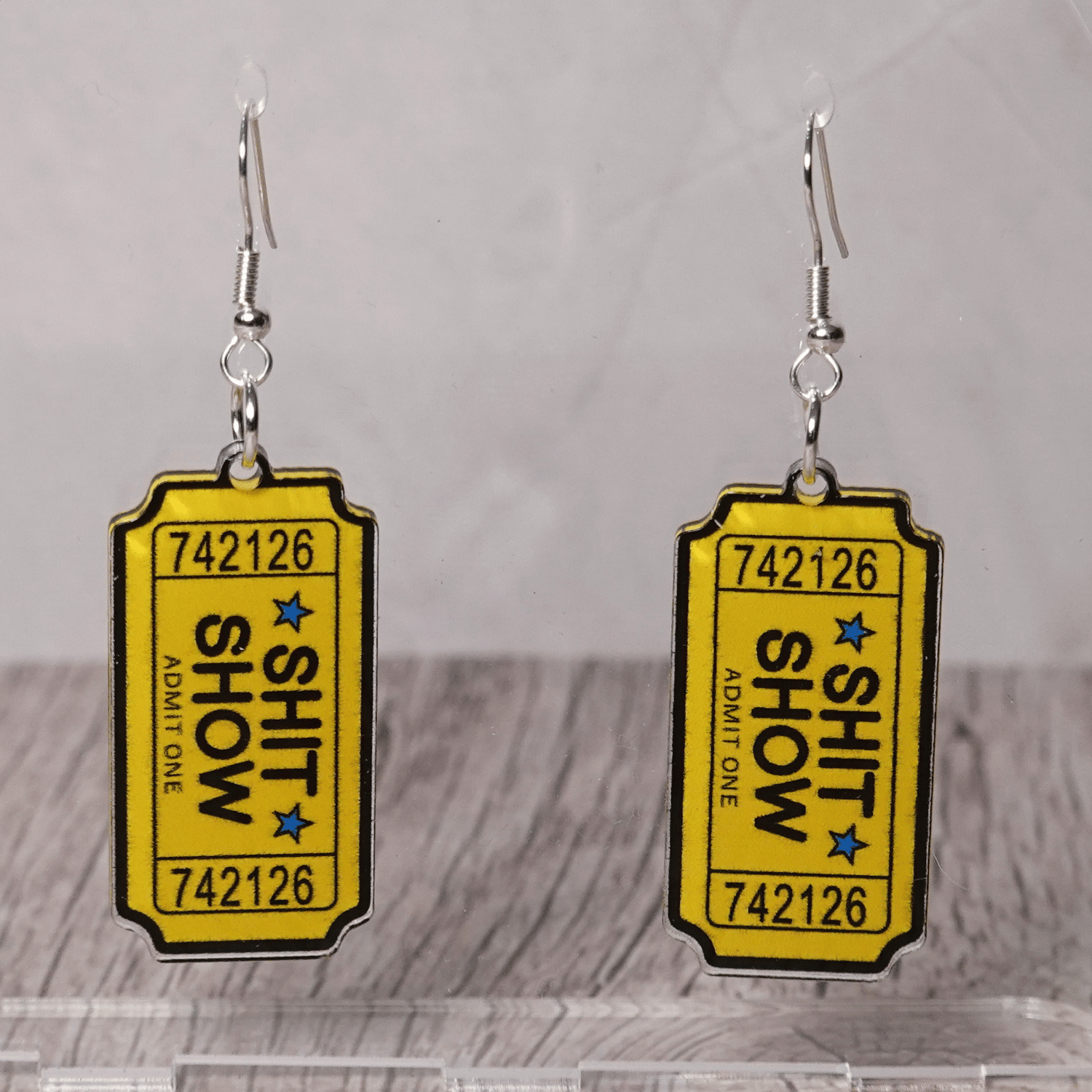 Acrylic dangle earrings designed like yellow admit-one tickets reading “Shit Show”