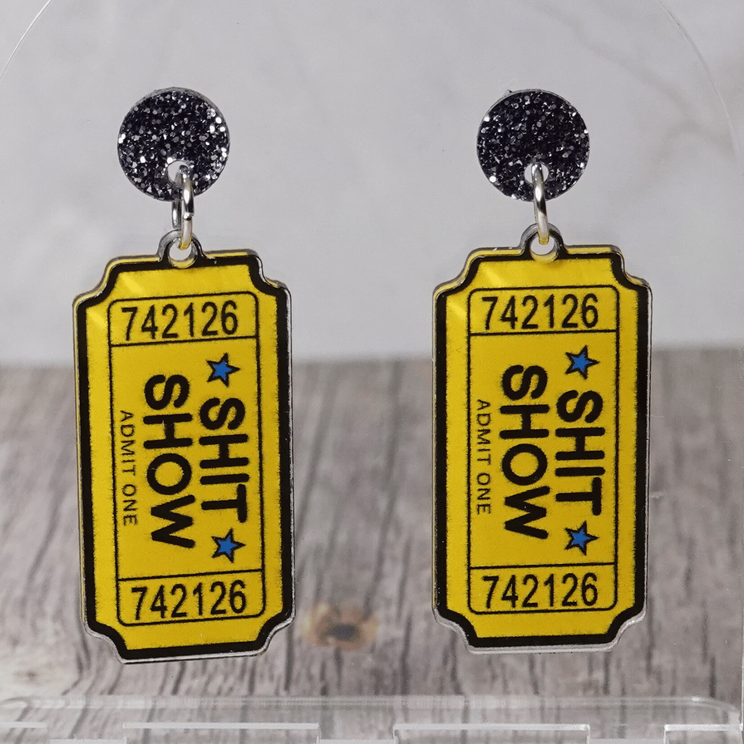 Acrylic dangle earrings designed like yellow admit-one tickets reading “Shit Show”