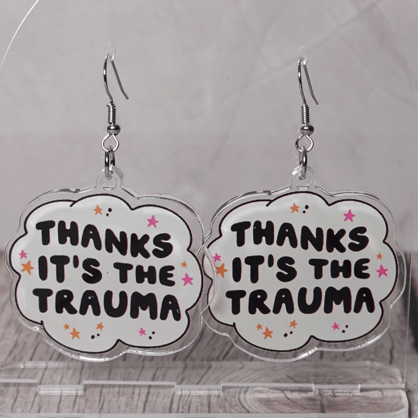 Clear acrylic dangle earrings with a speech bubble shape reading “Thanks it’s the trauma” with small star accents