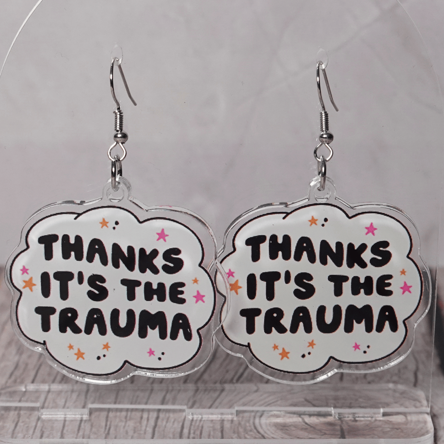 Clear acrylic dangle earrings with a speech bubble shape reading “Thanks it’s the trauma” with small star accents