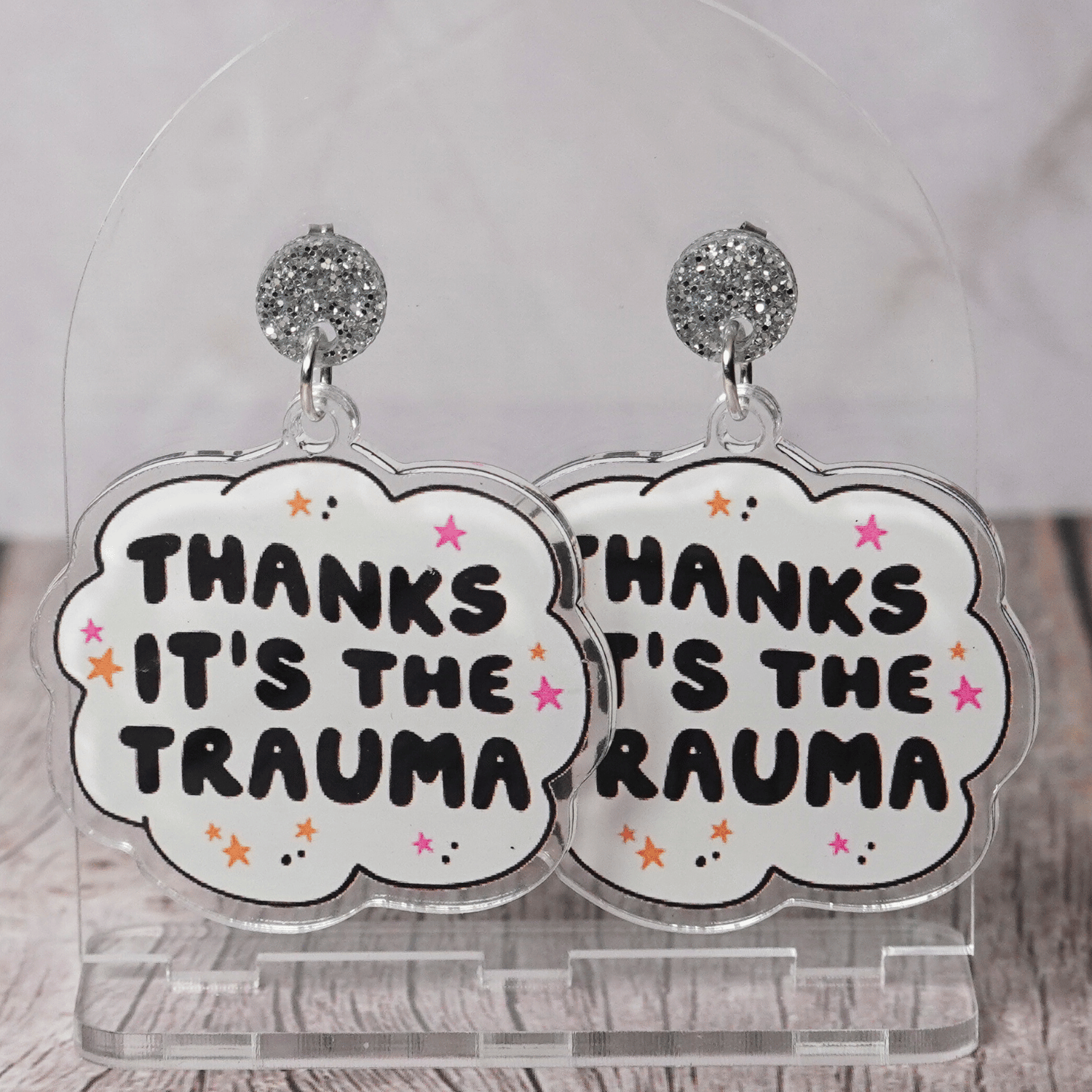 Clear acrylic dangle earrings with a speech bubble shape reading “Thanks it’s the trauma” with small star accents