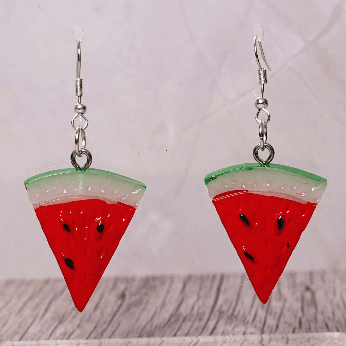 Watermelon slice dangle earrings shaped like red triangular watermelon pieces with green rind and black seed details on silver hooks.