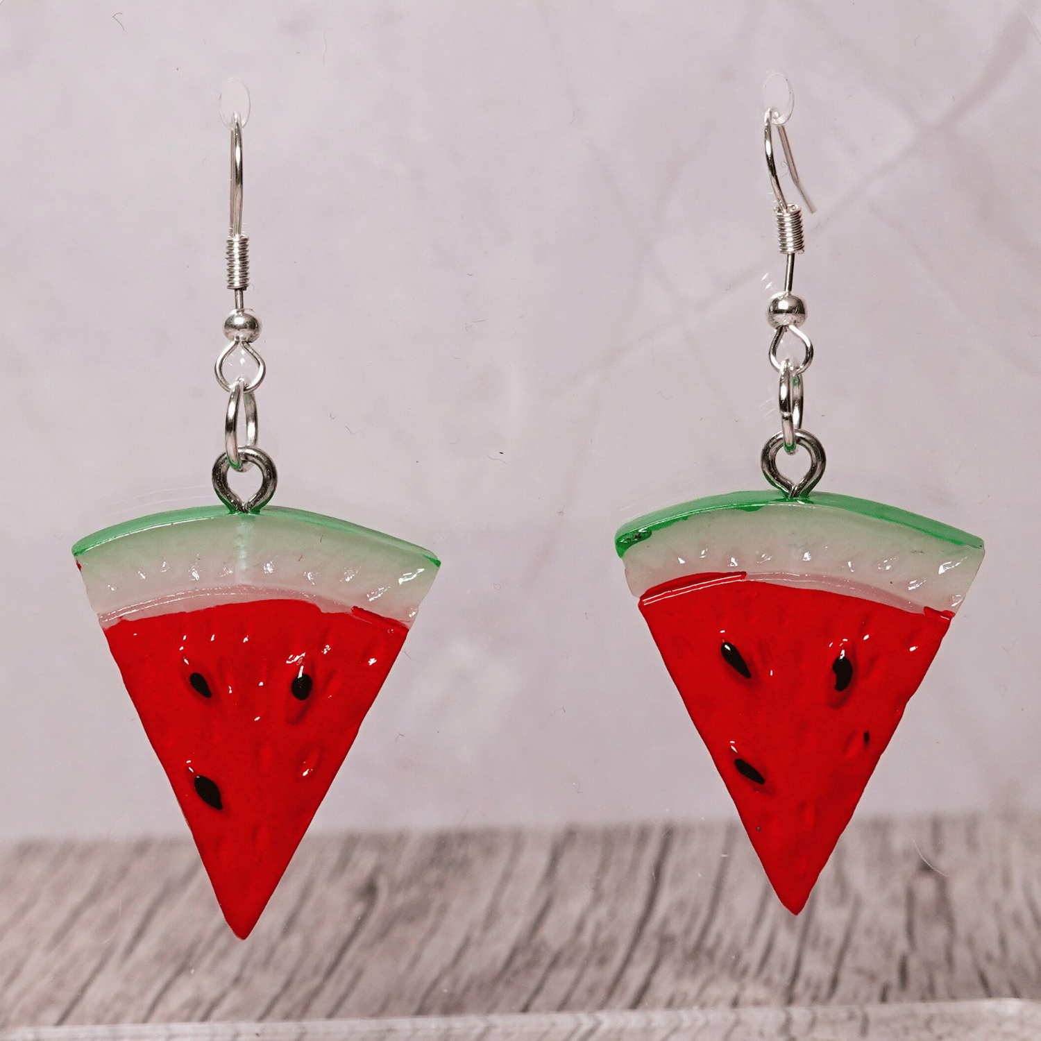Watermelon slice dangle earrings shaped like red triangular watermelon pieces with green rind and black seed details on silver hooks.