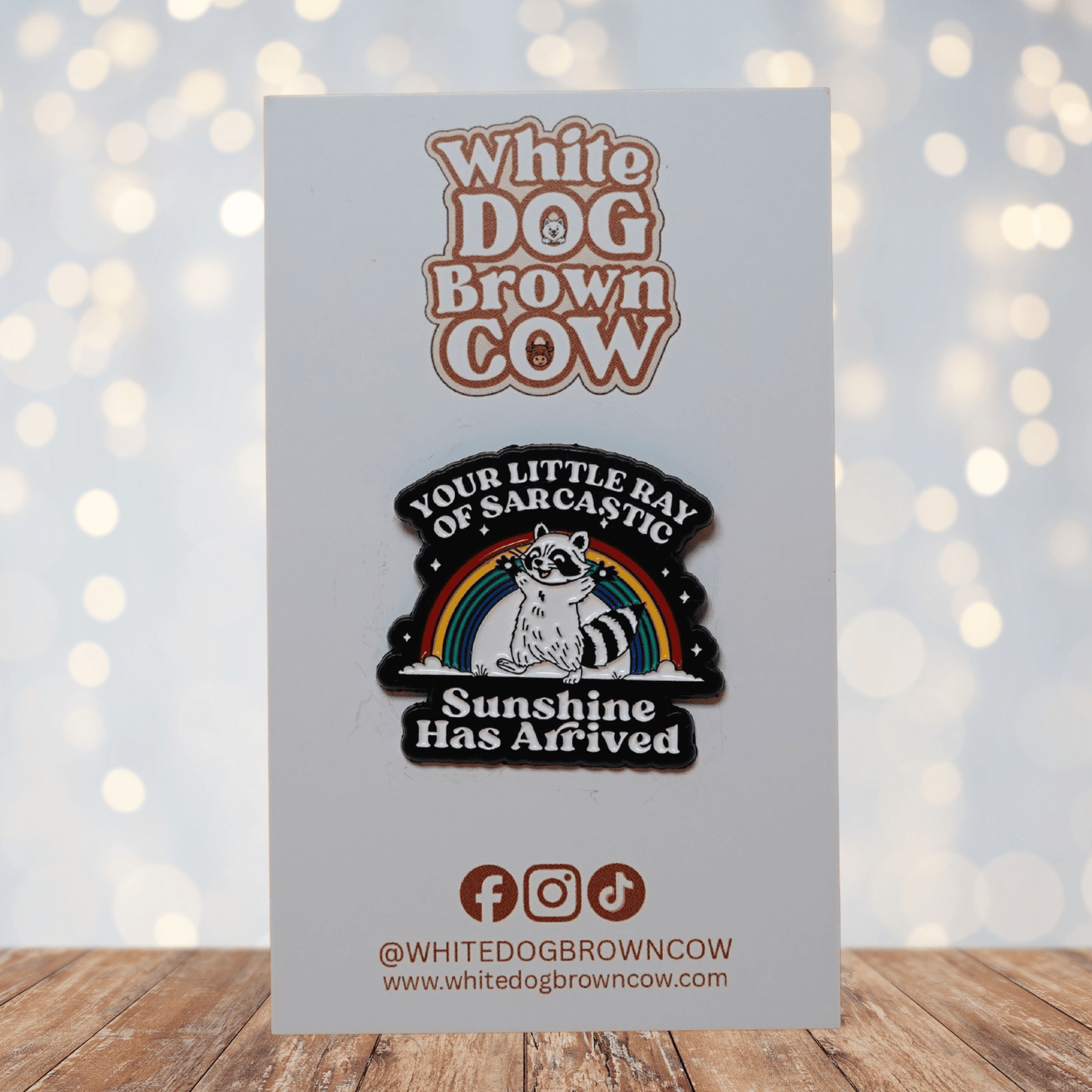 Enamel pin featuring a raccoon in front of a rainbow with the quote “Your little ray of sarcastic sunshine has arrived.”