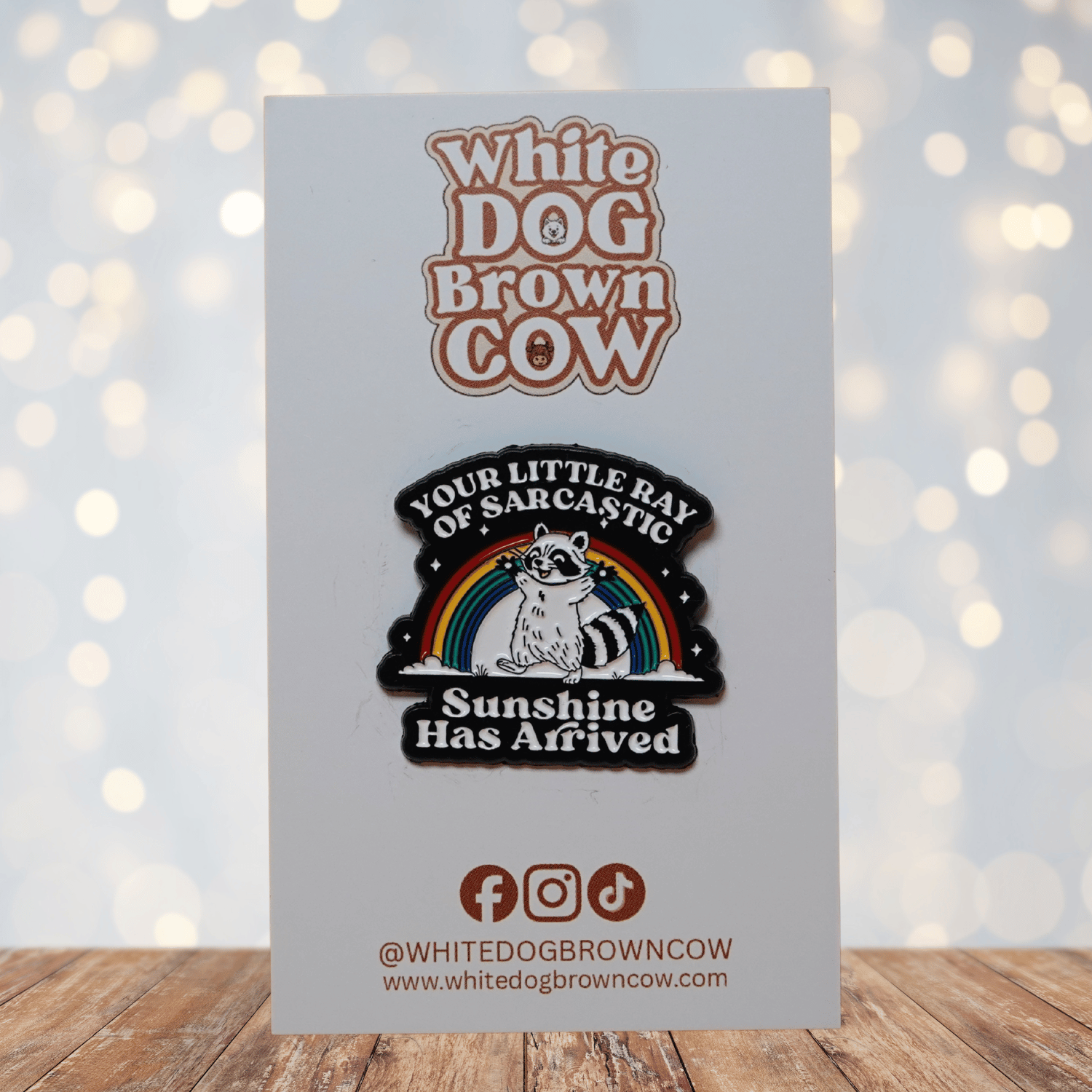 Enamel pin featuring a raccoon in front of a rainbow with the quote “Your little ray of sarcastic sunshine has arrived.”