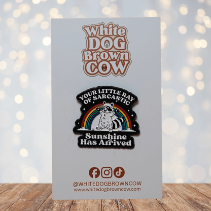 Enamel pin featuring a raccoon in front of a rainbow with the quote “Your little ray of sarcastic sunshine has arrived.”