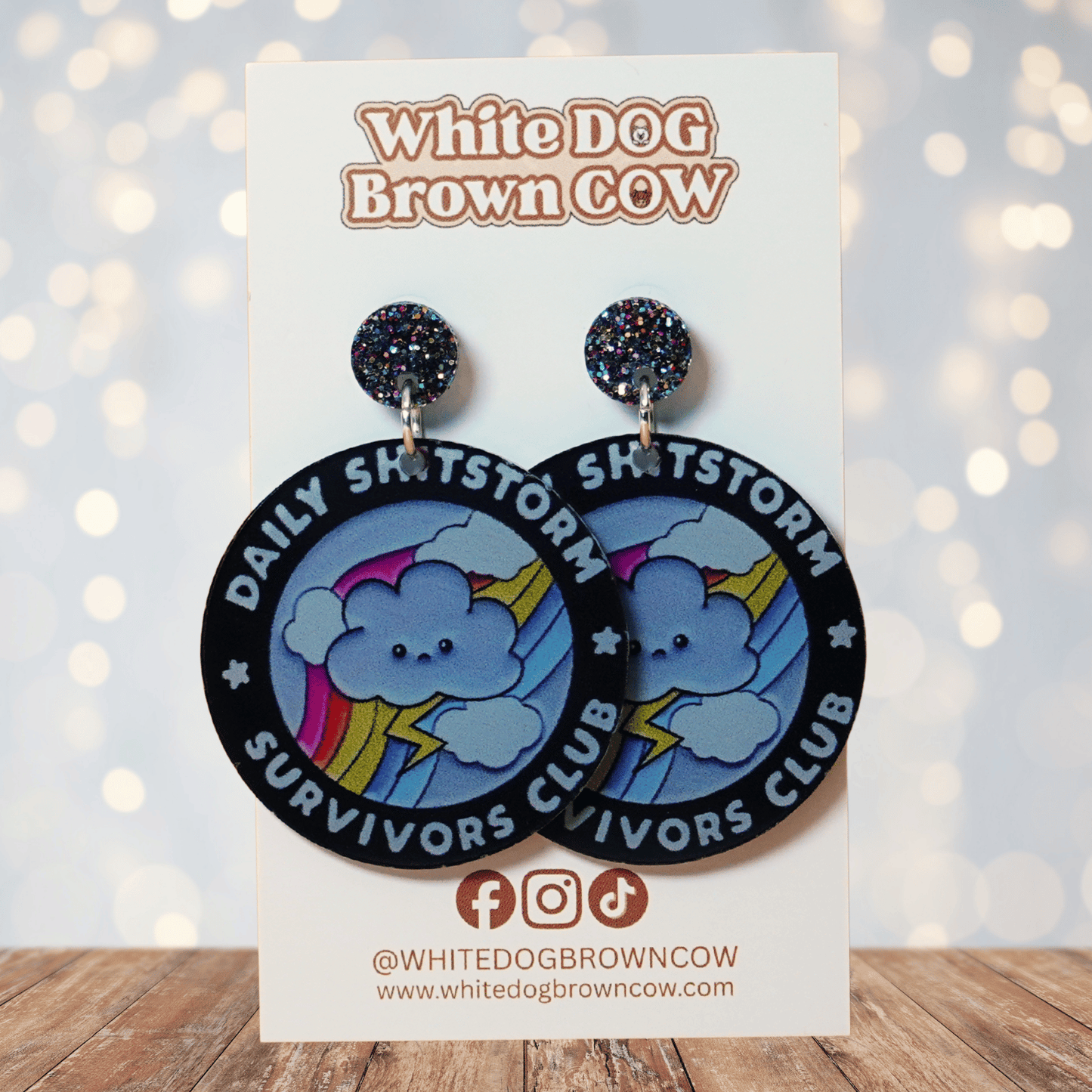 Round novelty dangle earrings reading “Daily Shitstorm Survivors Club” with a cute cloud, rainbow and lightning design