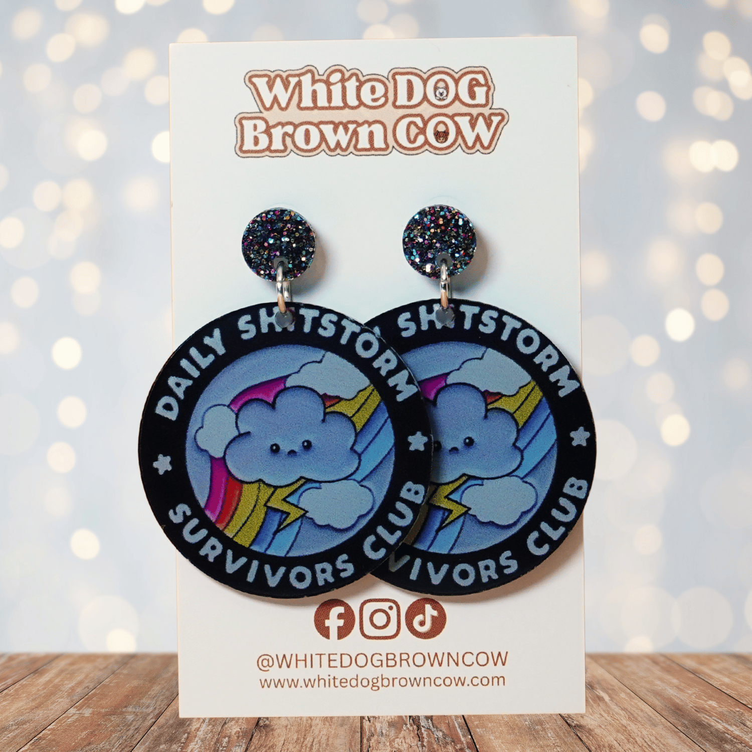 Round novelty dangle earrings reading “Daily Shitstorm Survivors Club” with a cute cloud, rainbow and lightning design