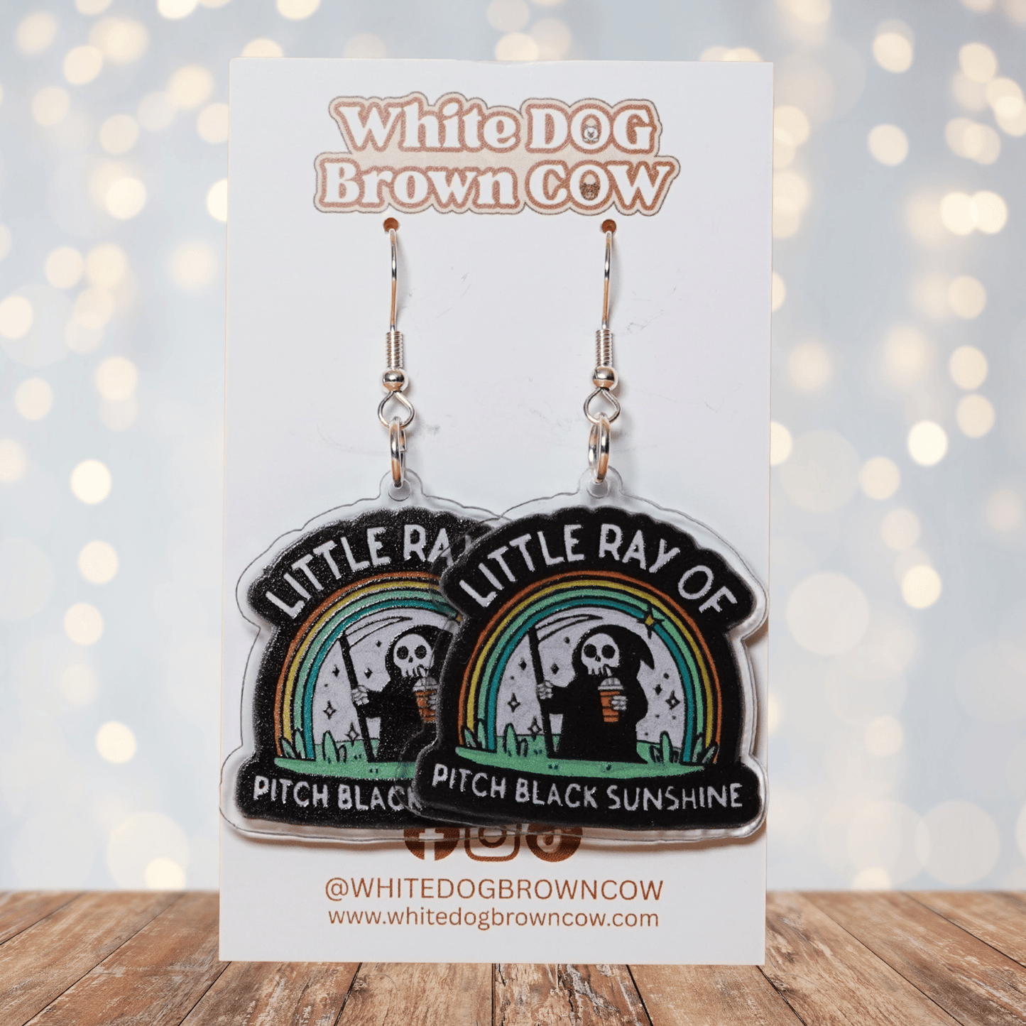 Novelty dangle earrings with a cartoon grim reaper holding a drink under a rainbow and the text “Little ray of pitch black sunshine”