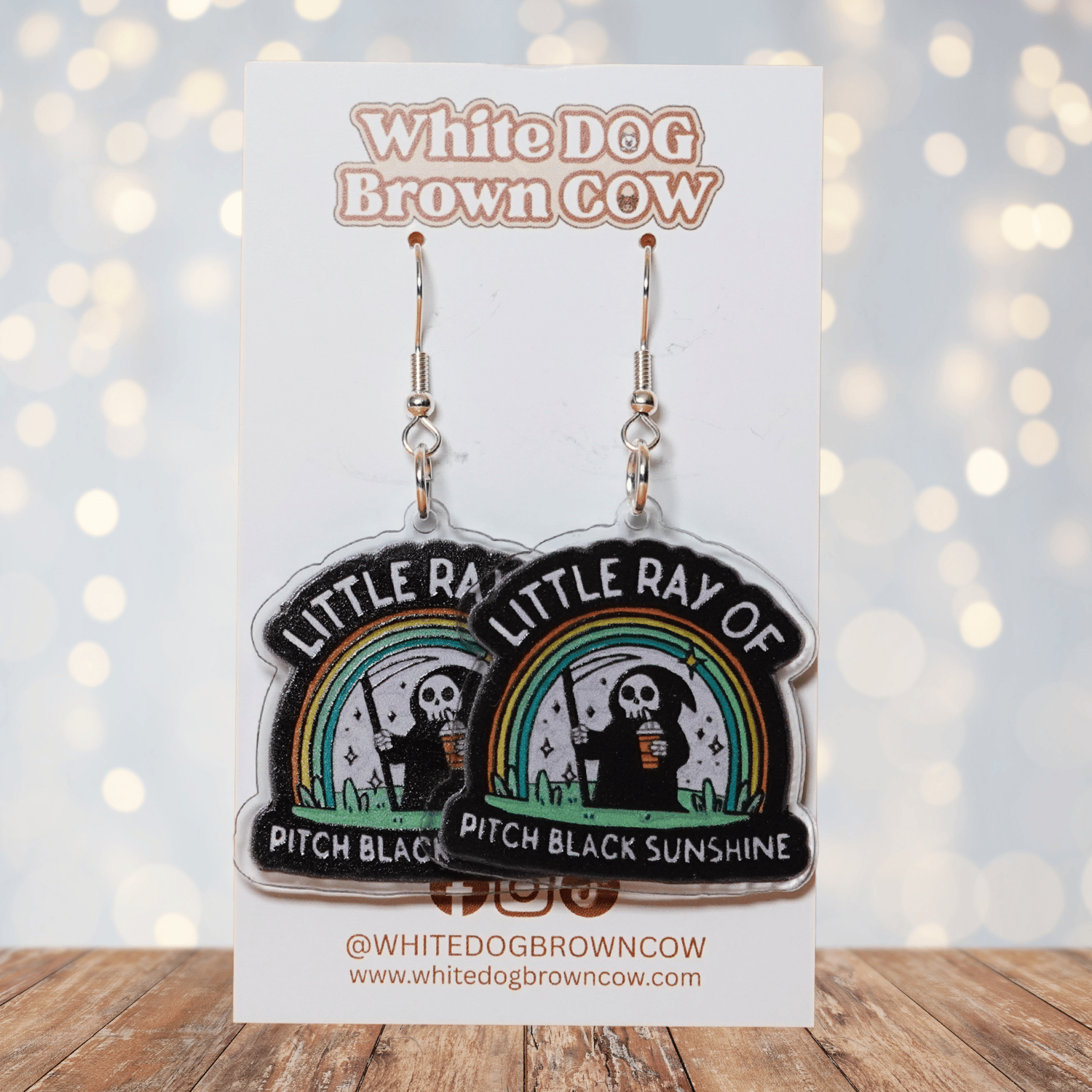 Novelty dangle earrings with a cartoon grim reaper holding a drink under a rainbow and the text “Little ray of pitch black sunshine”