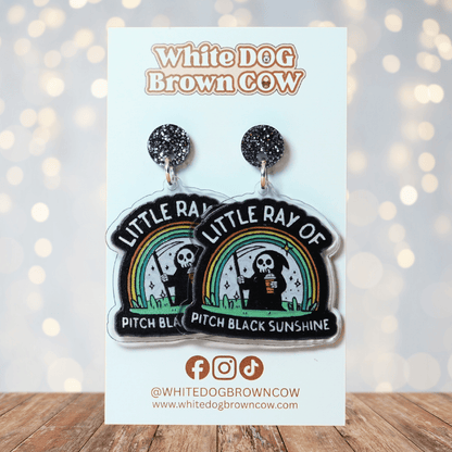 Novelty dangle earrings with a cartoon grim reaper holding a drink under a rainbow and the text “Little ray of pitch black sunshine”