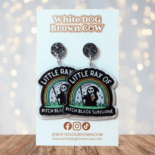 Novelty dangle earrings with a cartoon grim reaper holding a drink under a rainbow and the text “Little ray of pitch black sunshine”