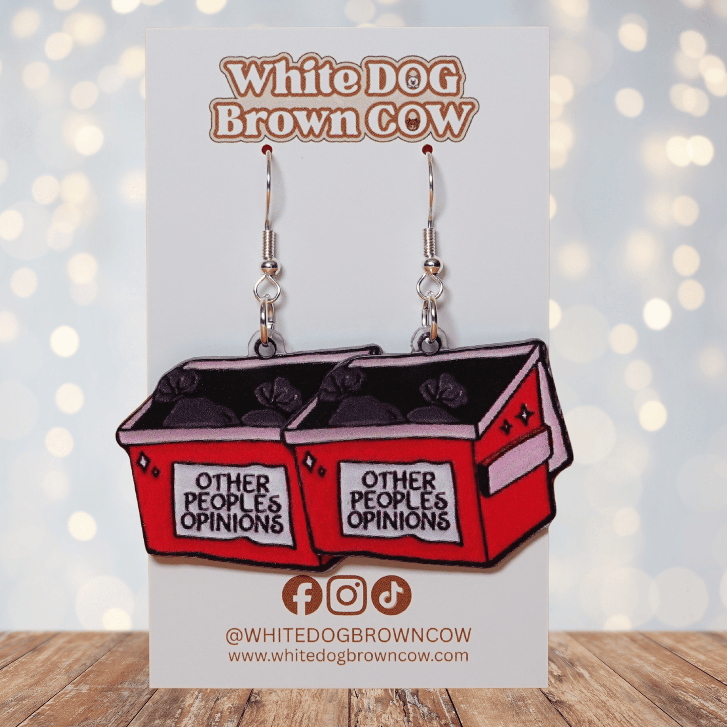 Acrylic dangle earrings shaped like a red dumpster labeled “Other People’s Opinions” with trash bags inside