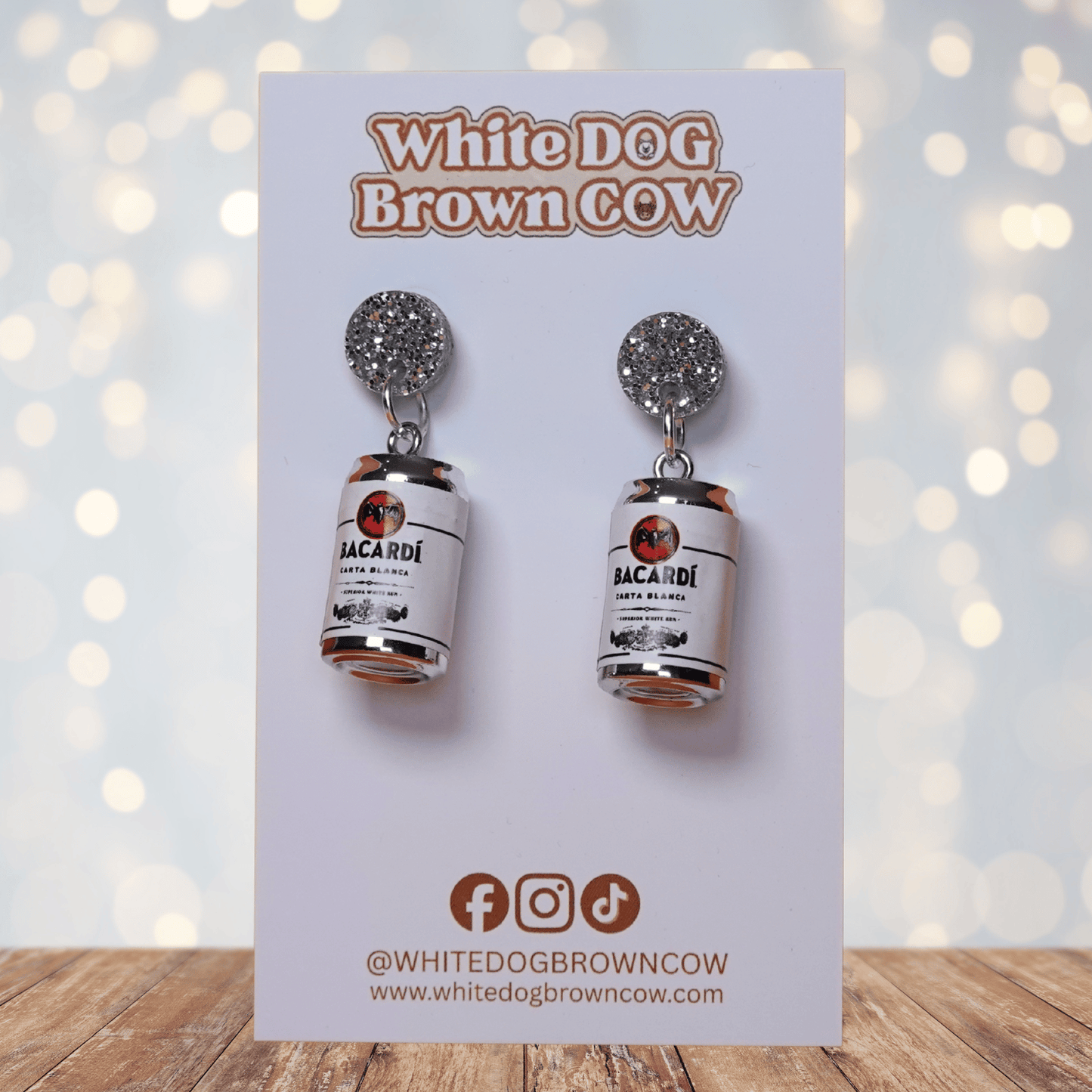 Novelty dangle earrings shaped like mini white rum cans with a printed label design and silver glitter studs.